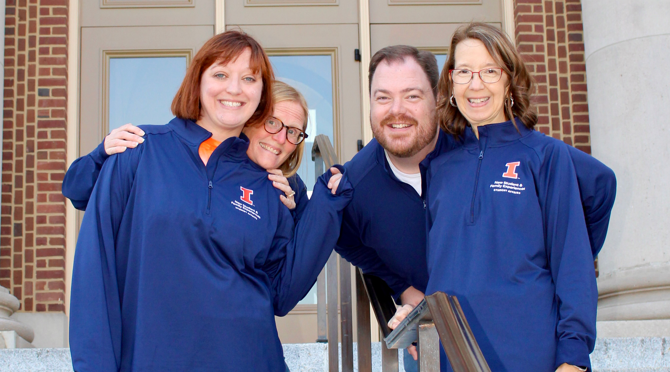 Four staff members smiling and embracing on steps in front of Foellinger Auditorium