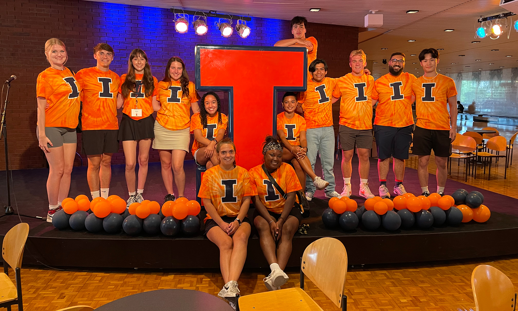 Group of 2023-2024 ITAs in orange shirts with blue Block I posing around large Block I cutout on stage.